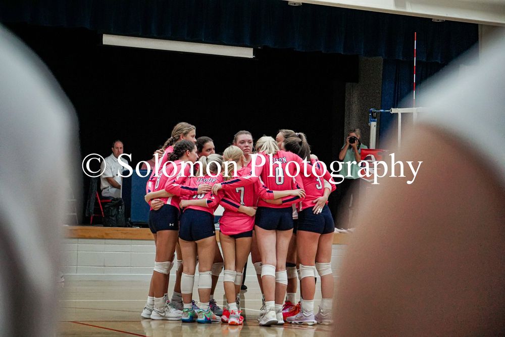 Girls volleyball team huddles together in pink and navy uniforms during an indoor match.