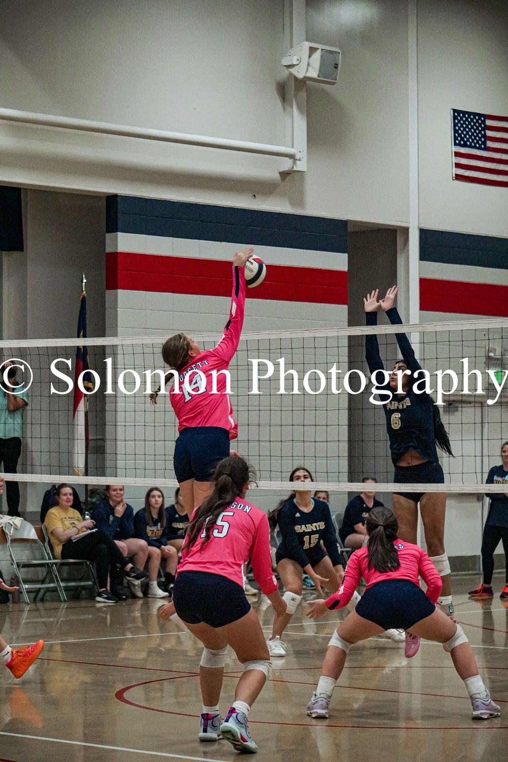 Girls volleyball player in pink jersey spikes ball over net during indoor high school match.