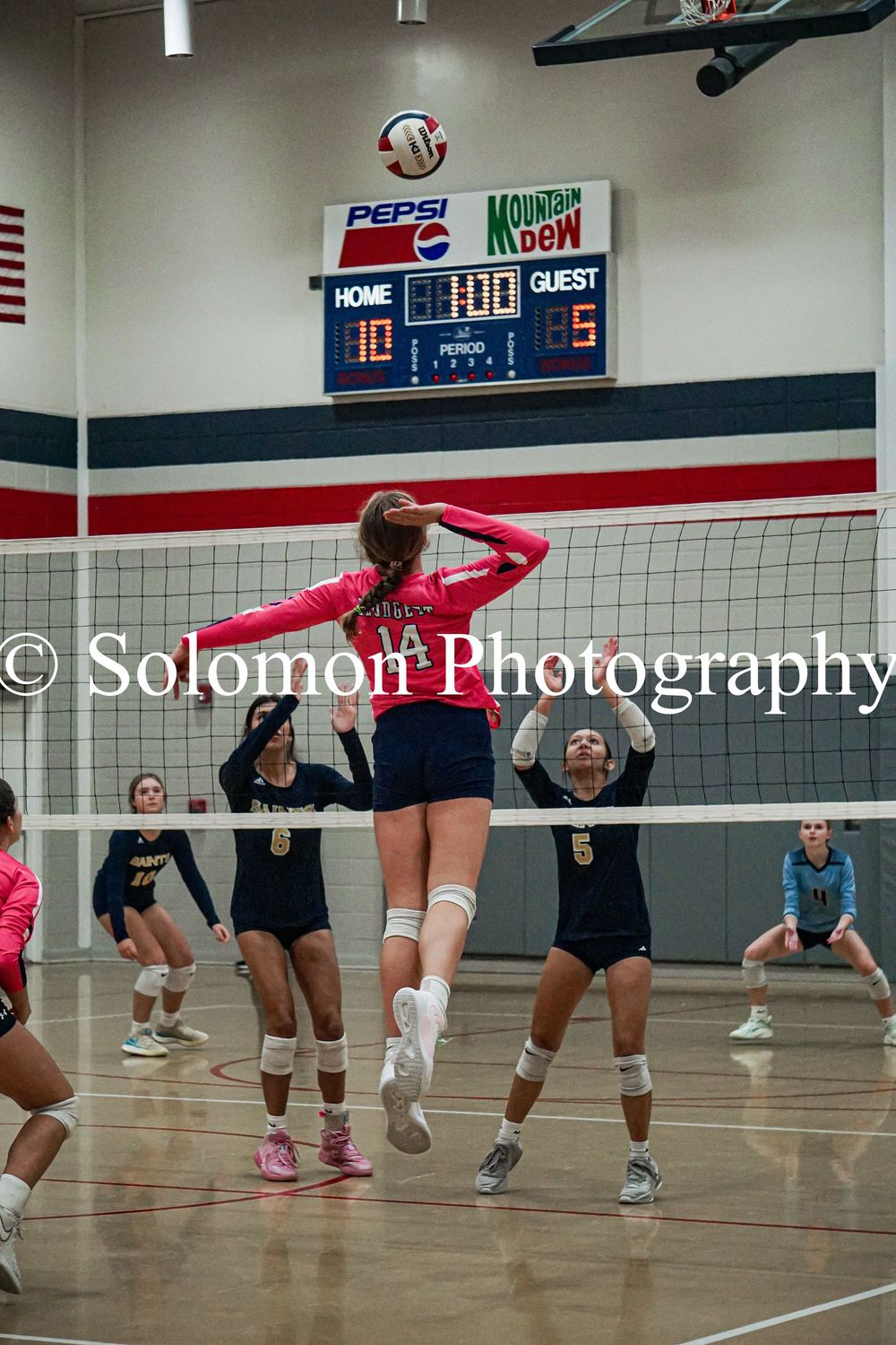 High school volleyball player jumps to spike ball over net during indoor match with score 10-5.