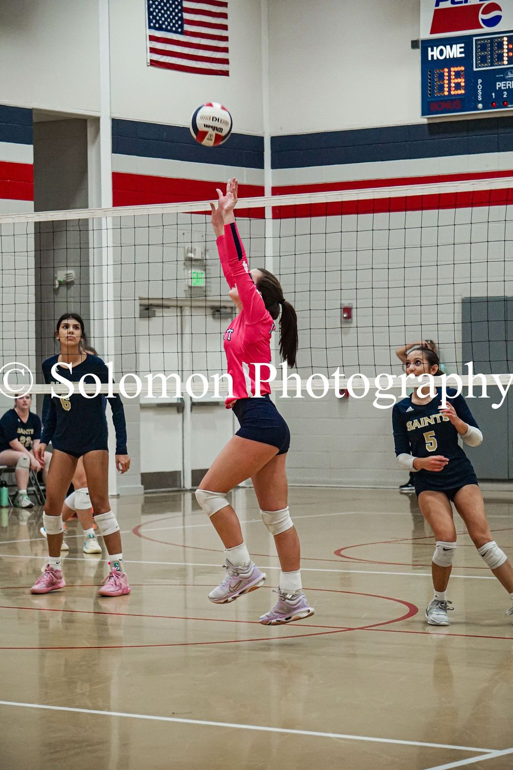 Female volleyball player in pink uniform sets ball during indoor high school game.