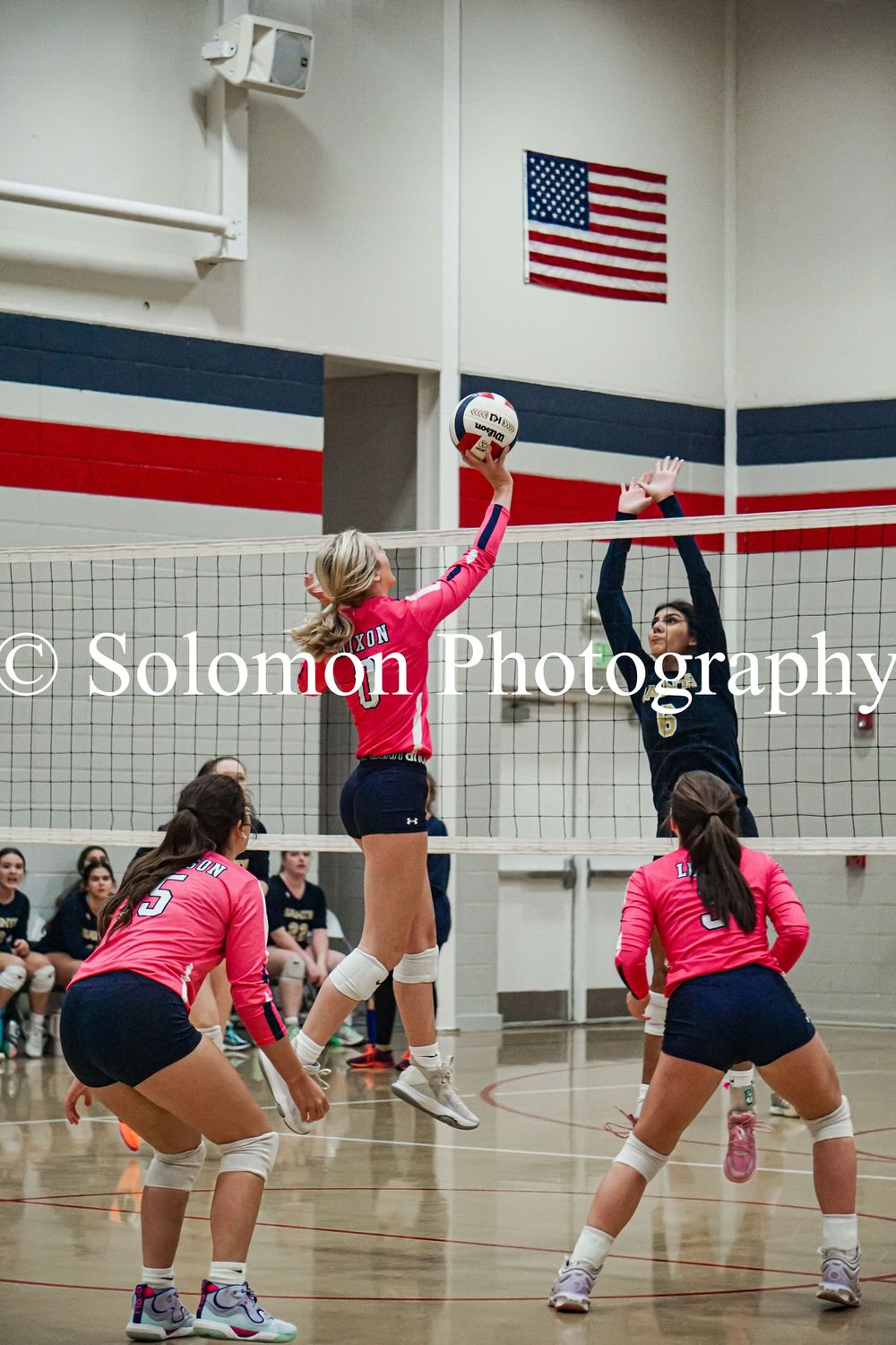 Girls volleyball player in red jersey tips ball over net against opponent during indoor match.