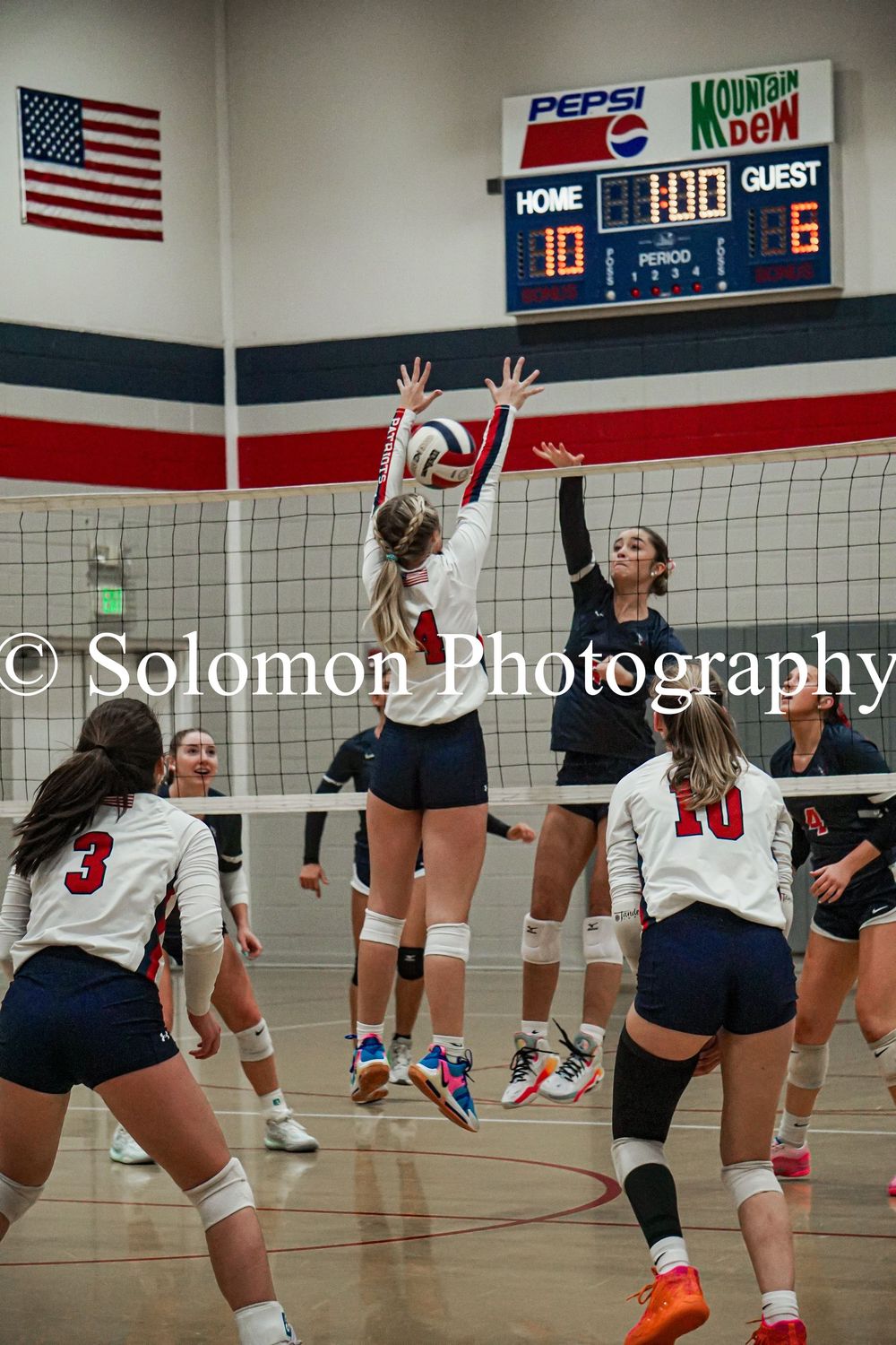 Two volleyball players block at the net during indoor high school game with score 10-6.