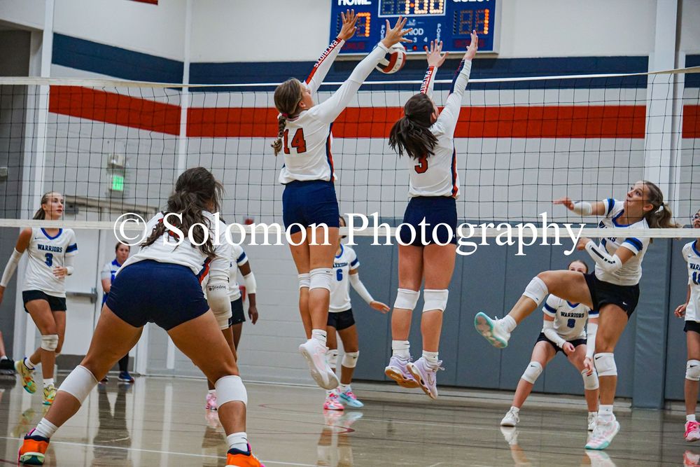 Two volleyball players in white uniforms jump to block spike during indoor high school game.