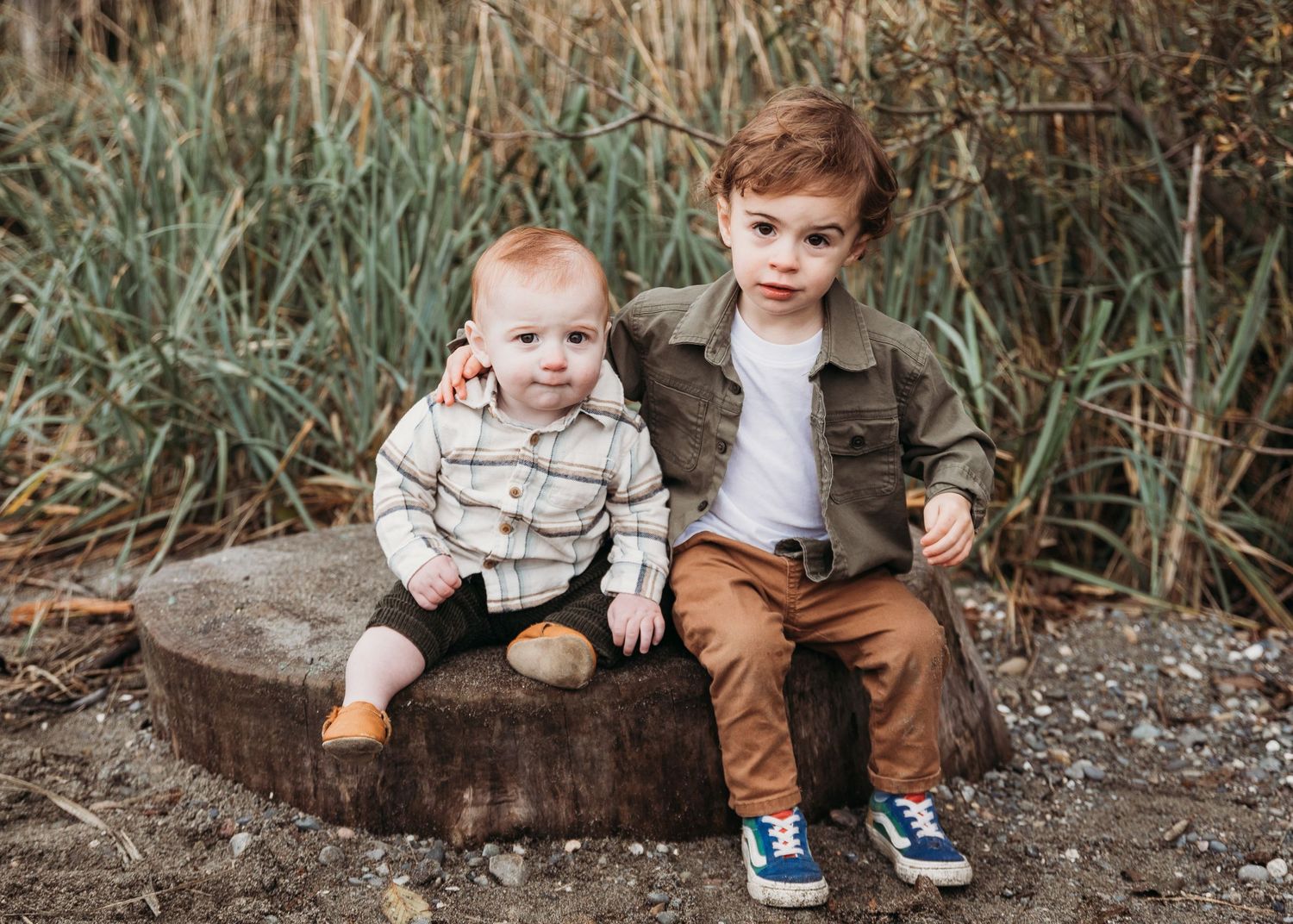 Two young children sit on a wooden log near tall grasses wearing casual autumn clothing.