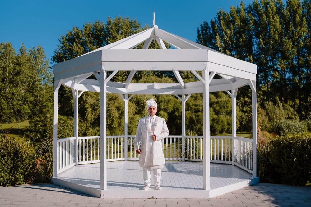 Groom in white sherwani standing in elegant white gazebo surrounded by lush green trees on sunny day.