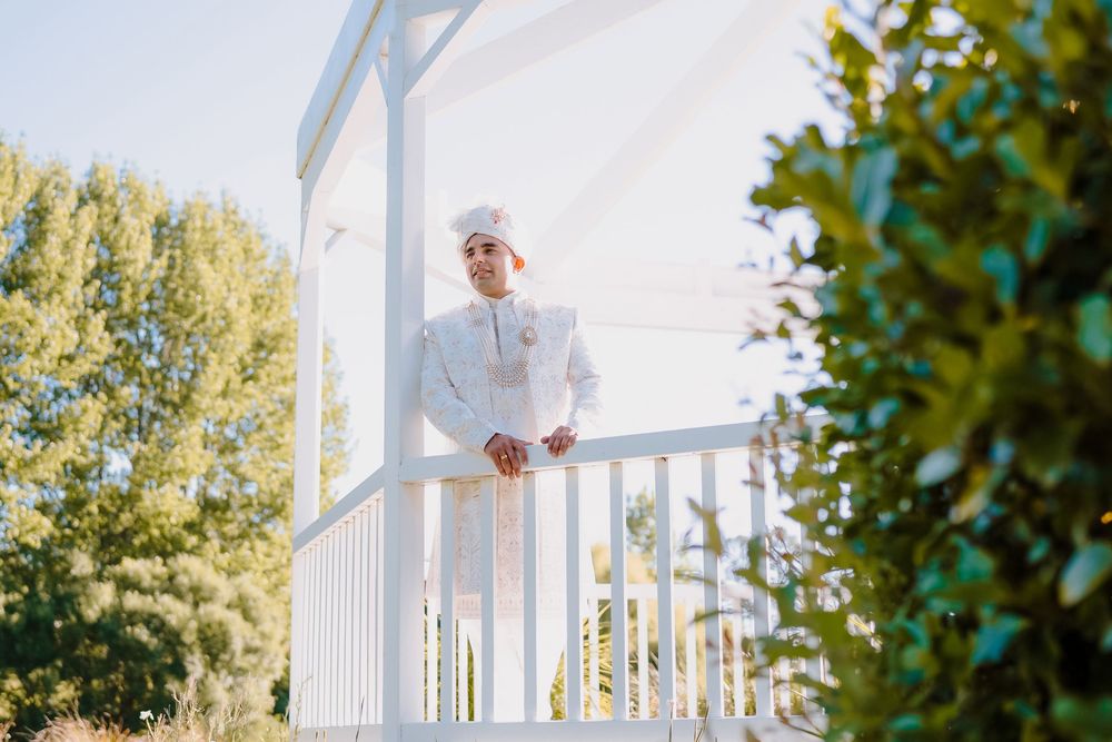 Groom in white traditional attire standing on a gazebo balcony surrounded by lush green trees on a sunny day.
