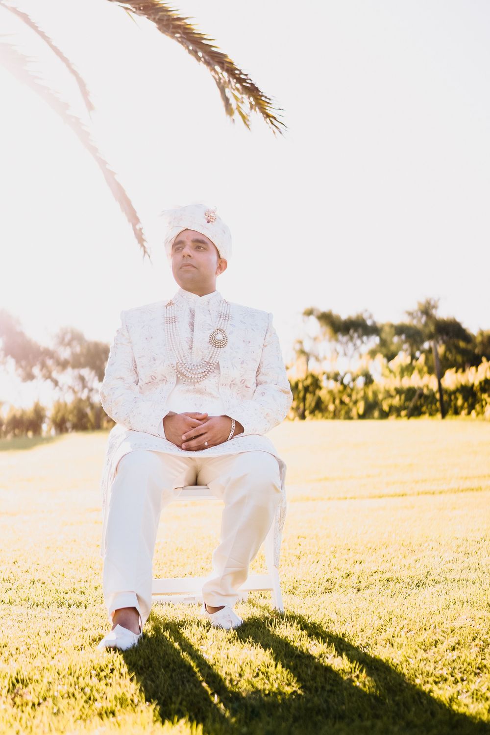 Indian groom in white sherwani and turban sits on chair outdoors during golden hour wedding ceremony.