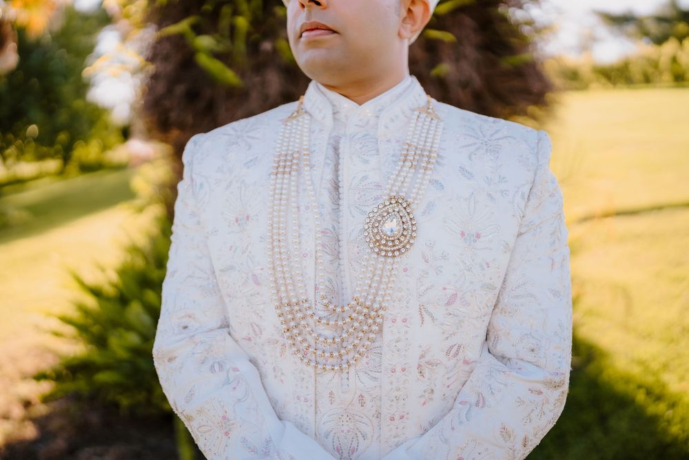 Groom in ornate white embroidered sherwani with layered pearl necklace at outdoor wedding ceremony.