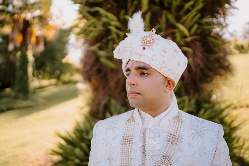 Indian groom in white embroidered sherwani and turban with floral brooch at outdoor wedding ceremony.