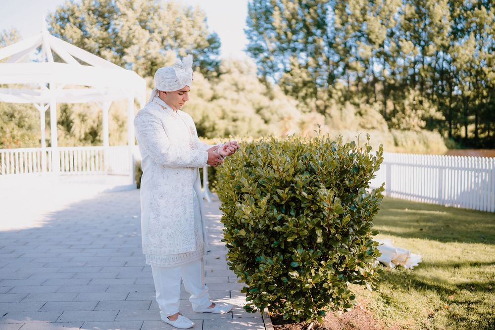 Indian groom in white embroidered sherwani adjusts boutonniere near garden gazebo.