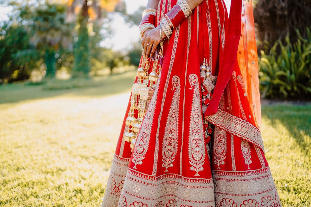 A bride in a stunning red and gold Indian lehenga with intricate embroidery stands on green grass.