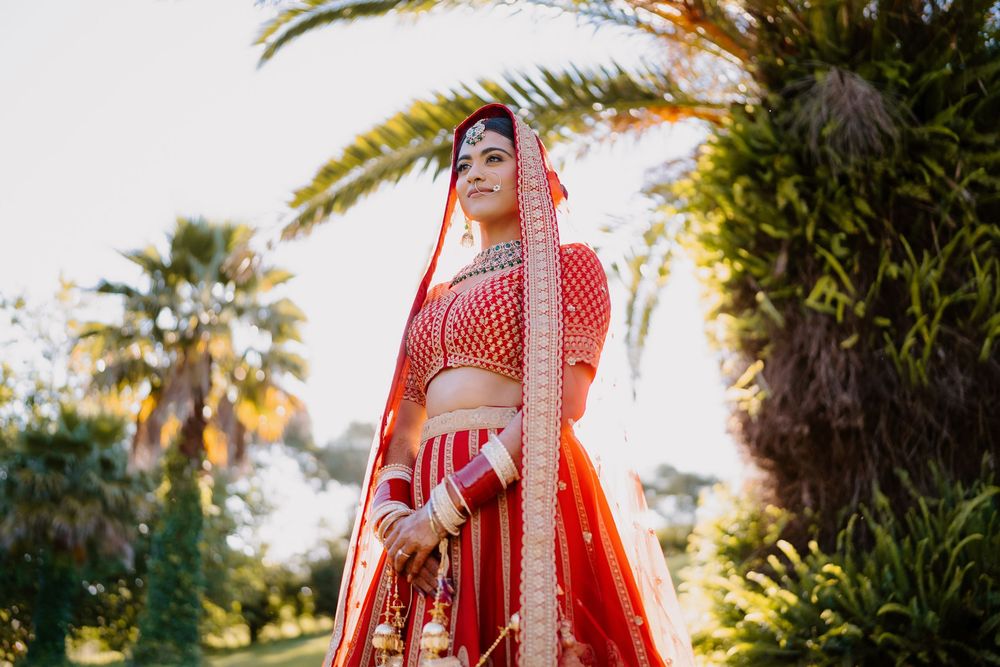 Indian bride in red lehenga with gold embroidery and dupatta poses outdoors near palm trees.