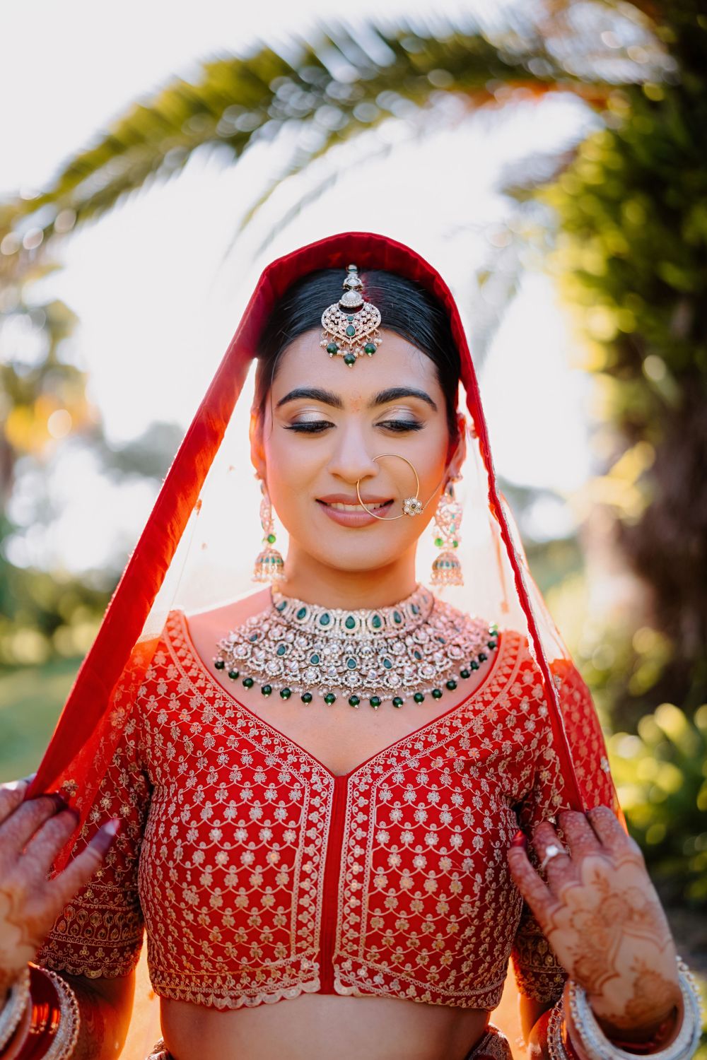 Indian bride in red lehenga with gold embroidery and emerald jewelry smiling downward on her wedding day.