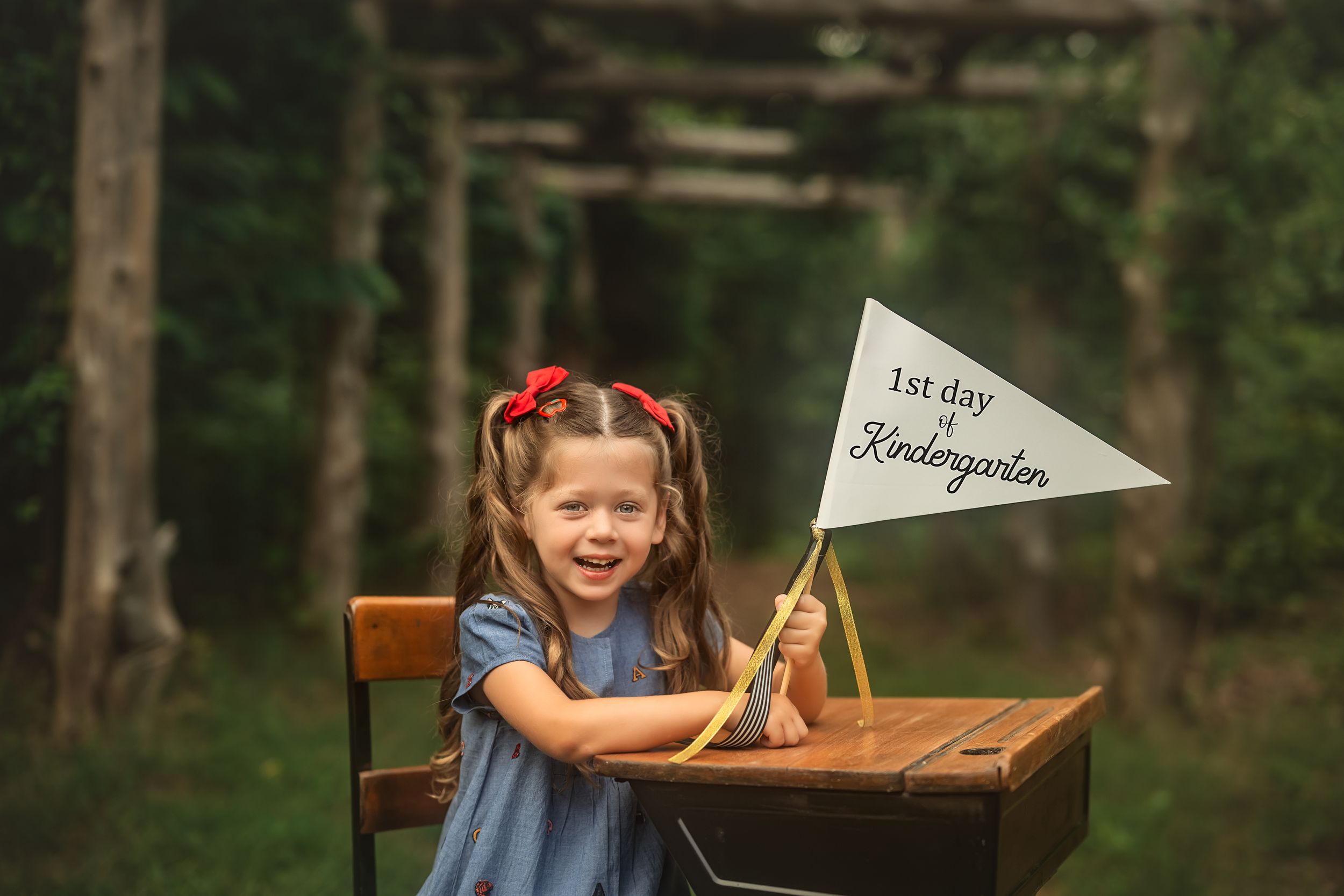 Student at antique desk holds 'First day of Kindergarten' flag in wooded outdoor setting.
