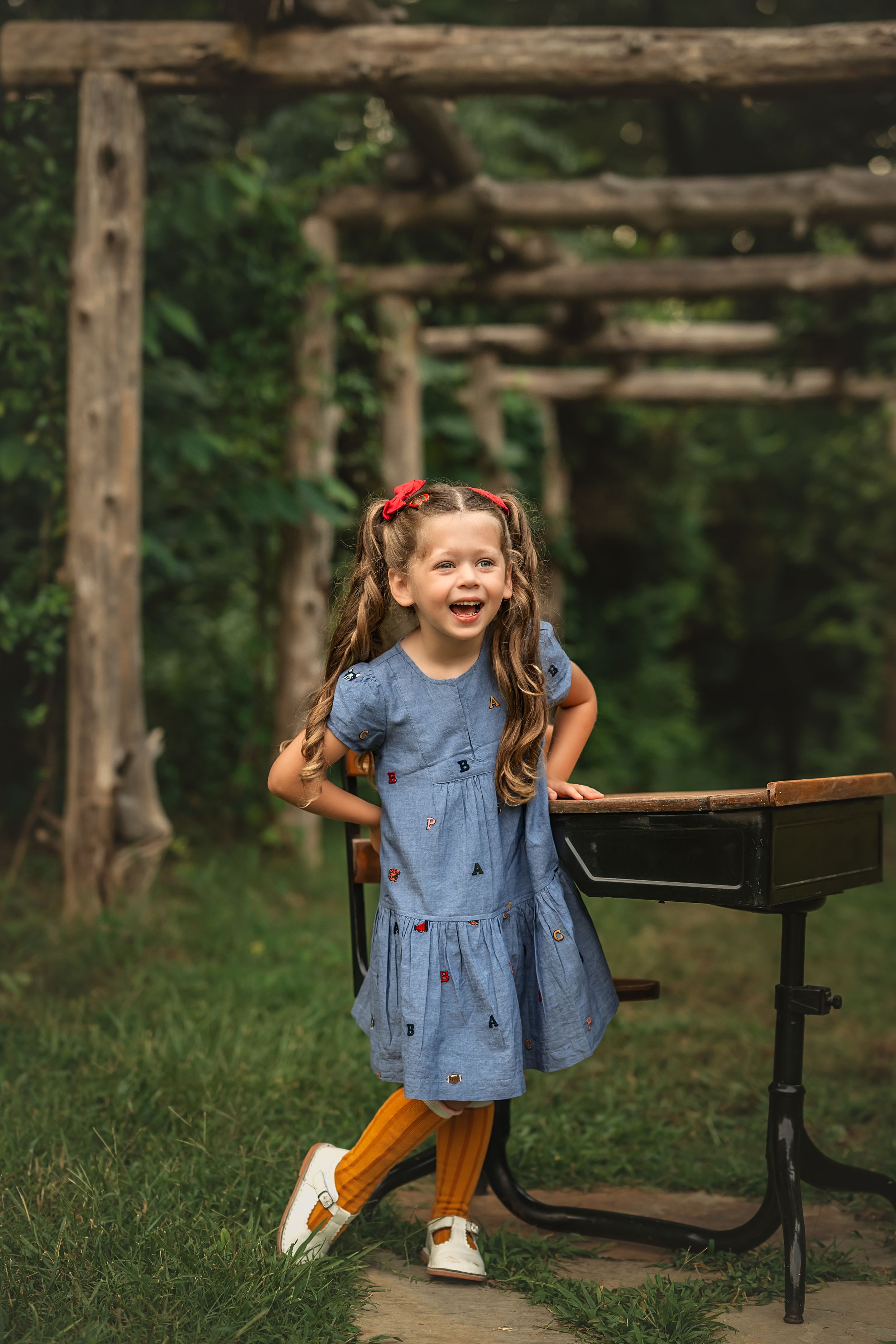 A joyful child in a blue denim dress and yellow pencil socks poses playfully next to an antique wooden school desk outdoors.