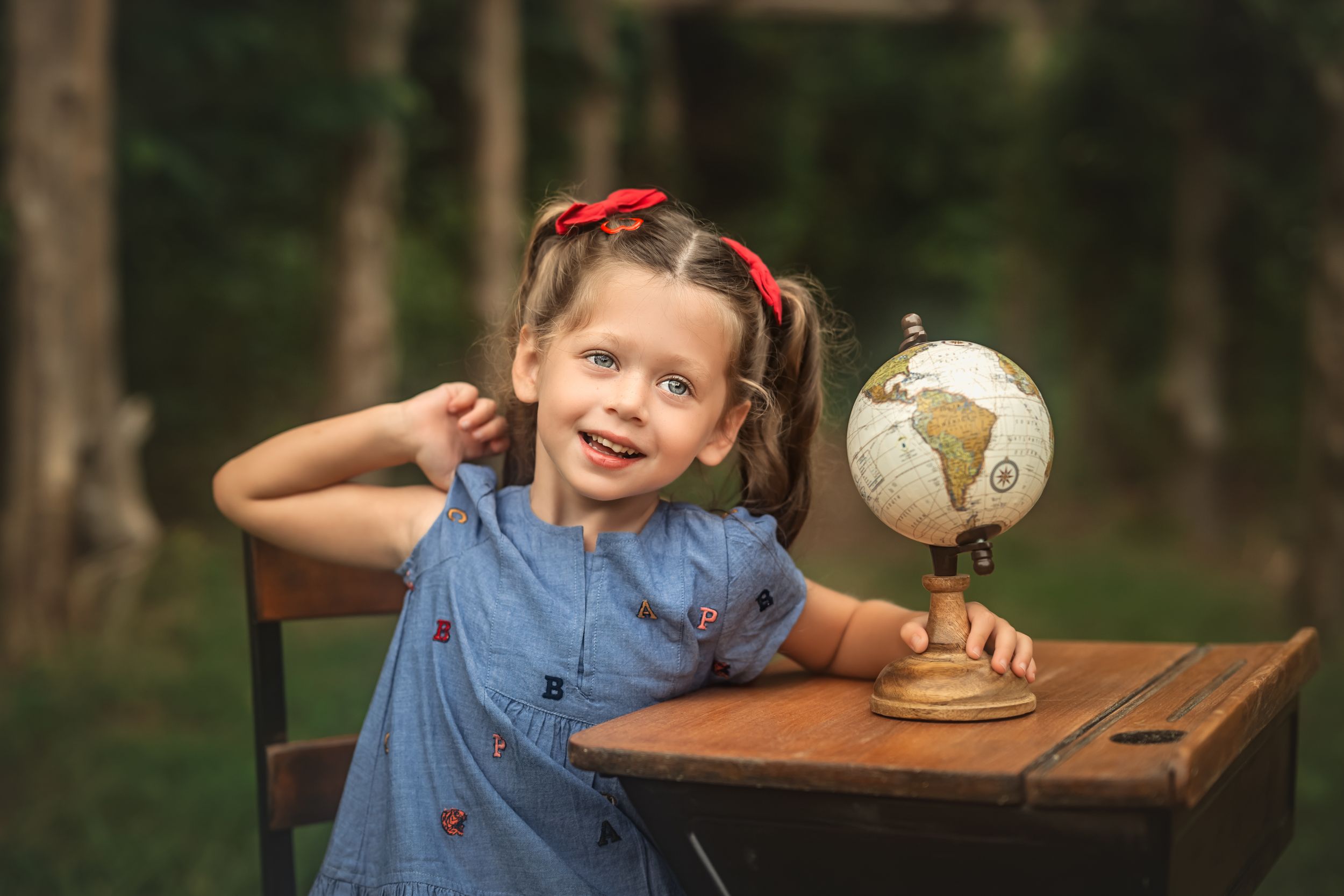 Child in blue dress and pigtails sits at wooden desk with vintage globe while posing in outdoor forest setting.