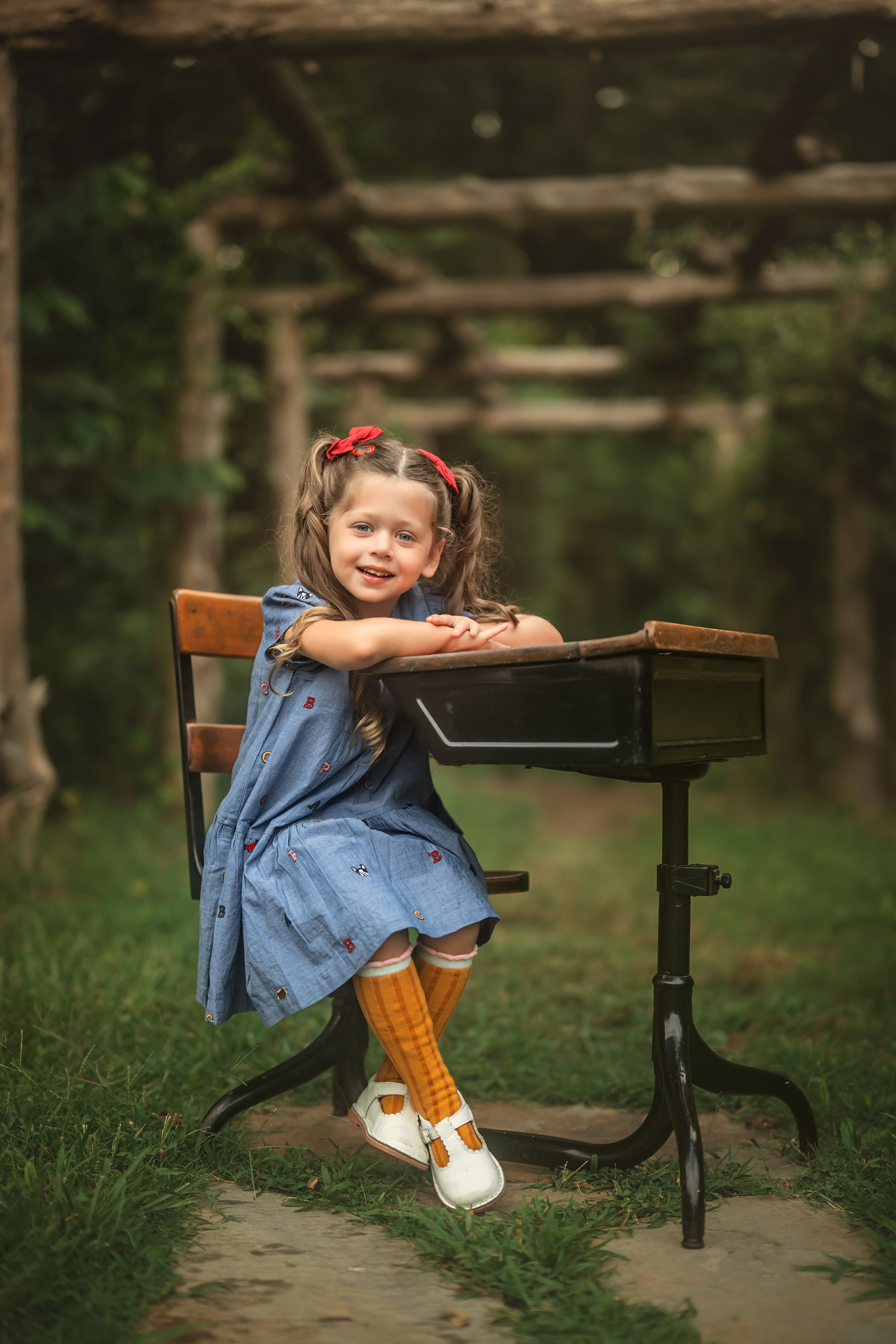A young girl in a blue dress and pigtails sits at a vintage school desk in a lush garden setting.