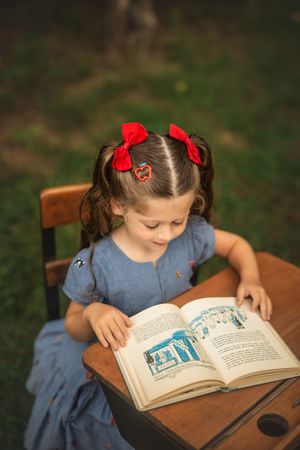 A child reads an open storybook on an antique wooden school desk in a garden setting.