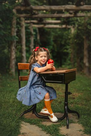 A nostalgic scene with a vintage school desk and little girl in a blue dress against a wooded backdrop.