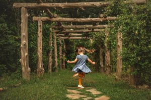 A carefree child in blue dress skips through a rustic wooden pergola in a lush garden.