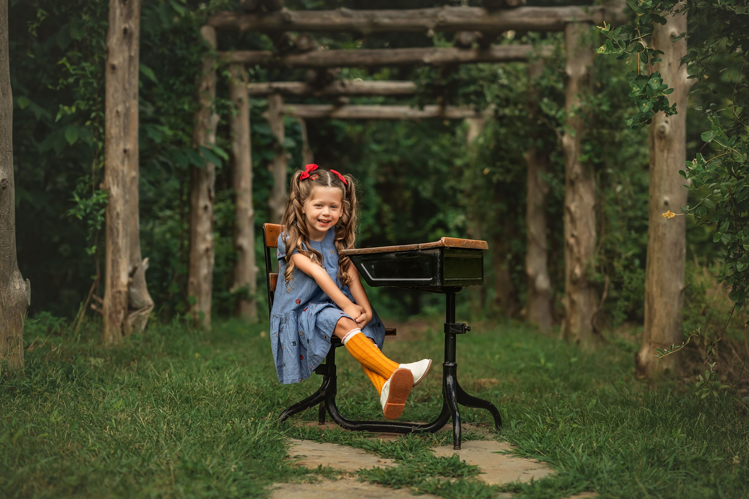 A child sits at an antique school desk for back-to-school photos on a path through trees with sunlight filtering through the foliage.