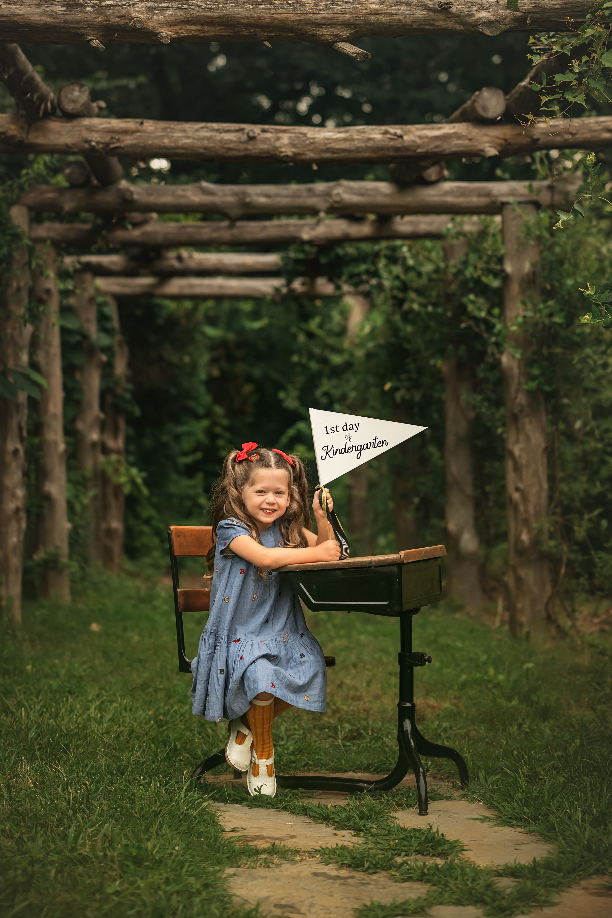 A girl sits at an old-fashioned student desk with wooden seat on a grass path surrounded by rustic wooden posts.