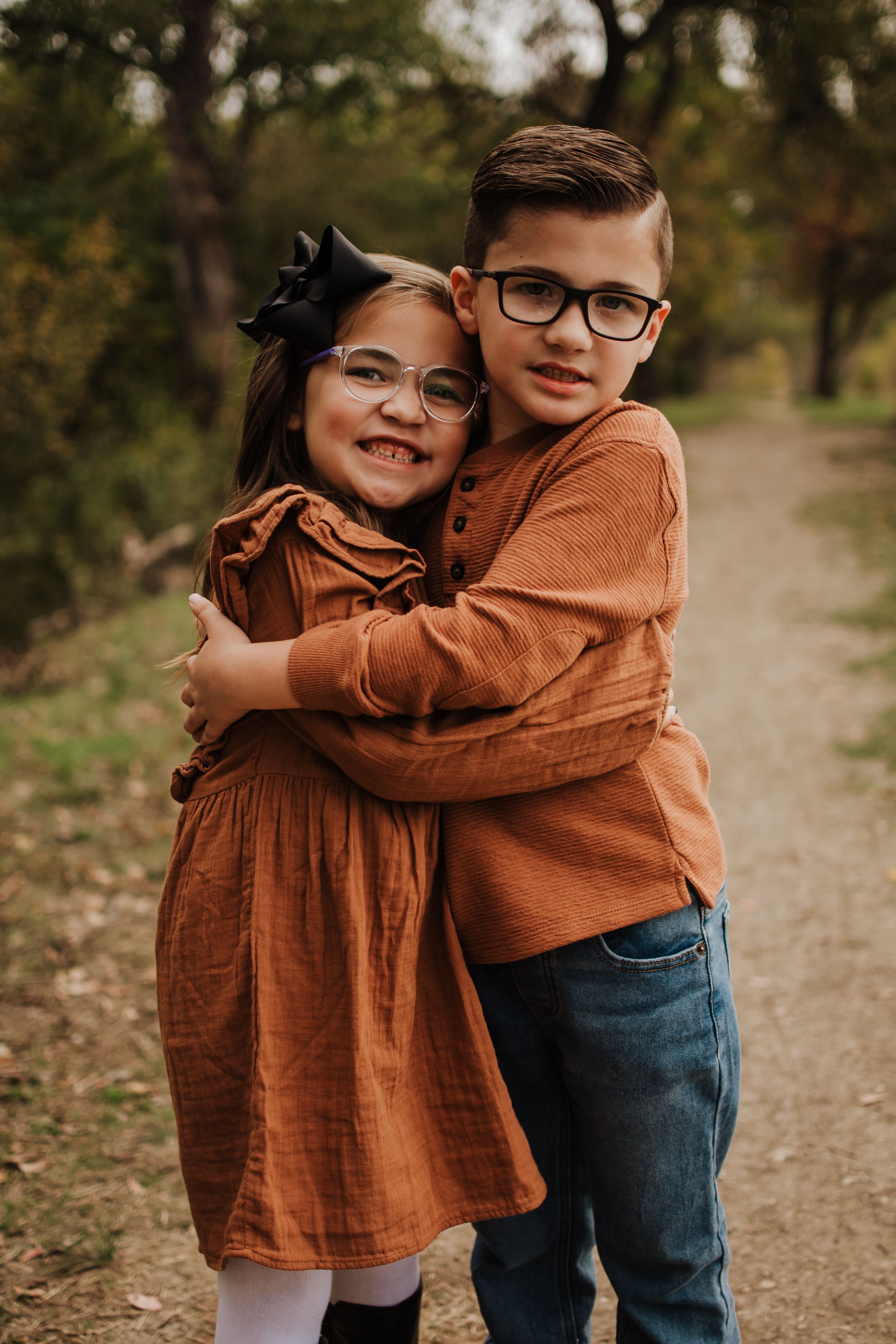 Two children in matching orange outfits share a warm embrace on a fall path.