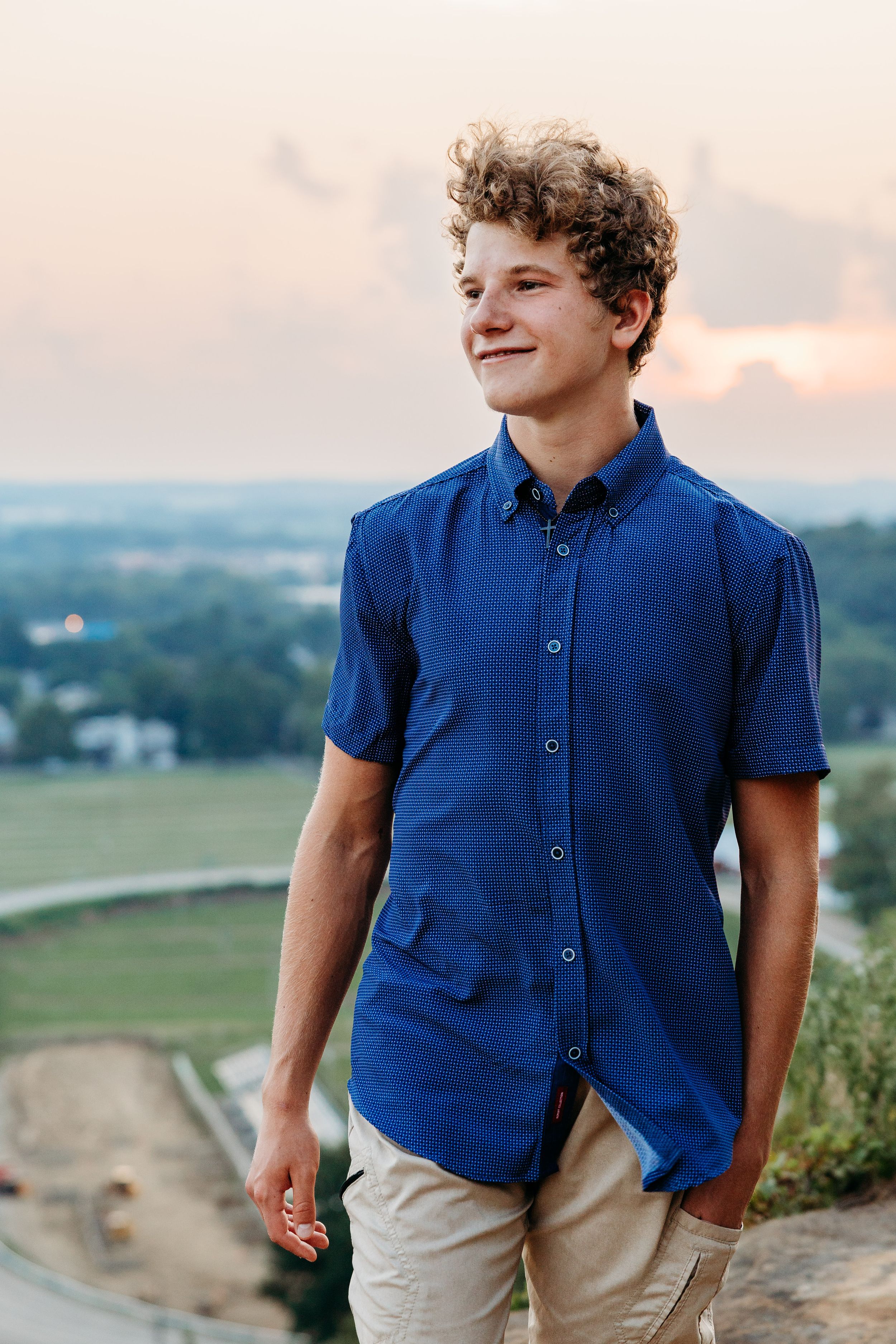 A person in a navy blue button-down shirt and khaki pants stands on a scenic overlook at sunset.