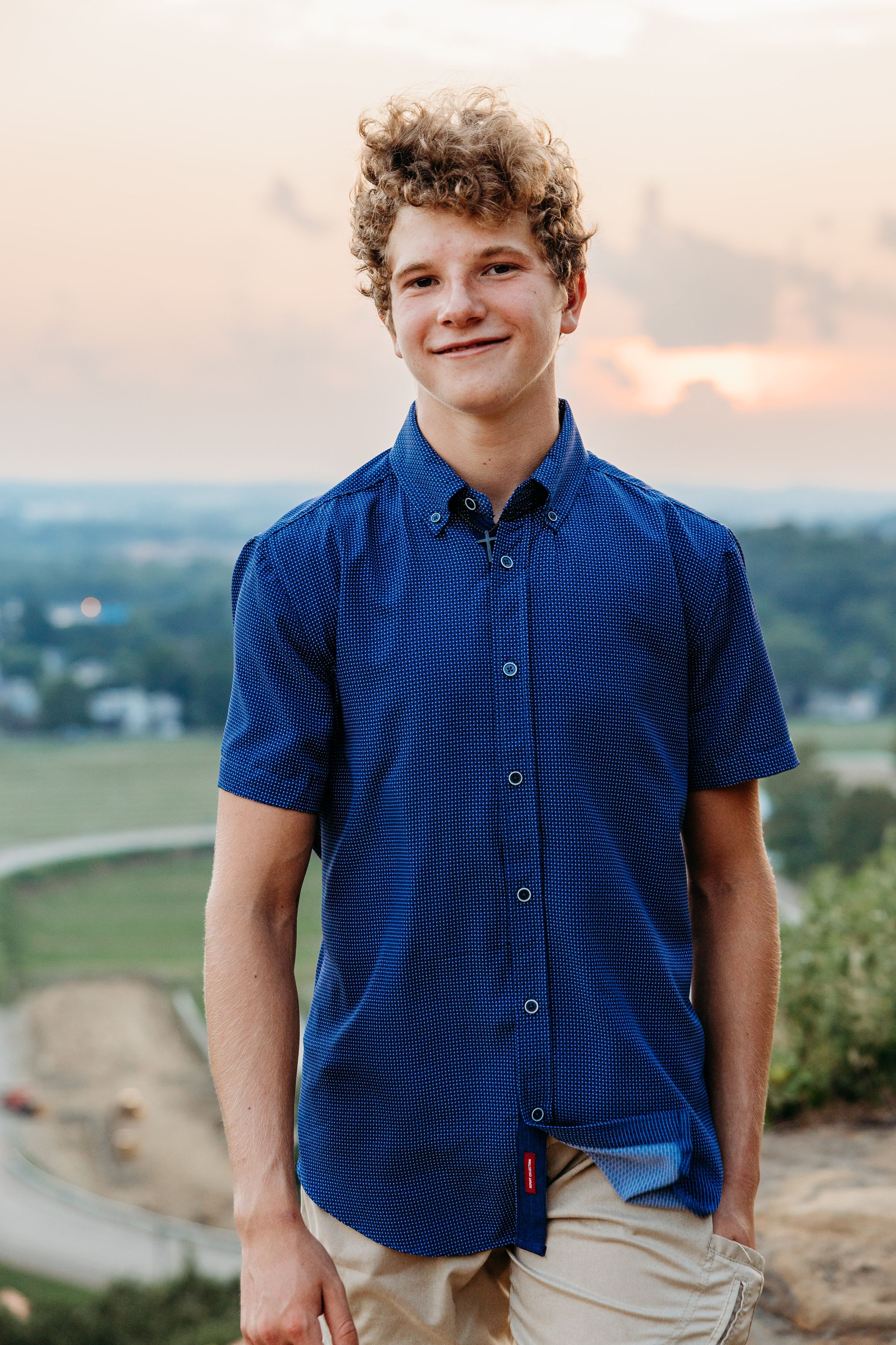 Person in blue short-sleeve button down shirt stands outdoors against sunset sky and scenic overlook.