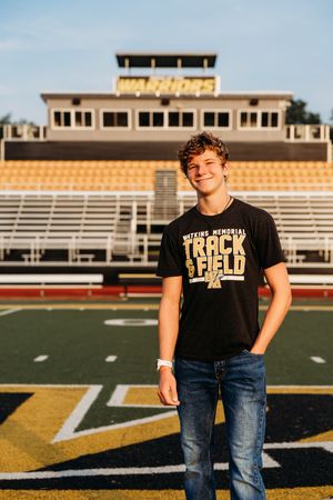 Track and field athlete poses in stadium wearing black team t-shirt and jeans at sunset.