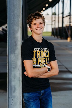 Student in black track and field t-shirt and jeans leans against column with crossed arms and watches nearby.