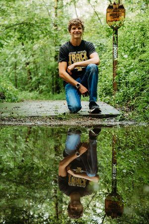 A reflection in still water creates a mirror image of someone kneeling on a wooden boardwalk in a lush green forest.