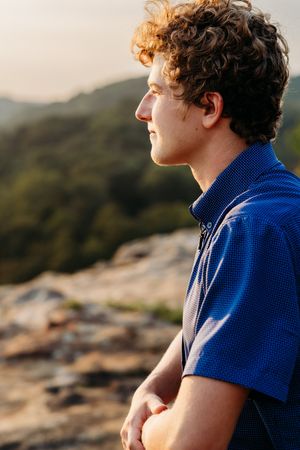 Person in blue polo shirt looking thoughtfully at sunset from rocky mountain viewpoint.