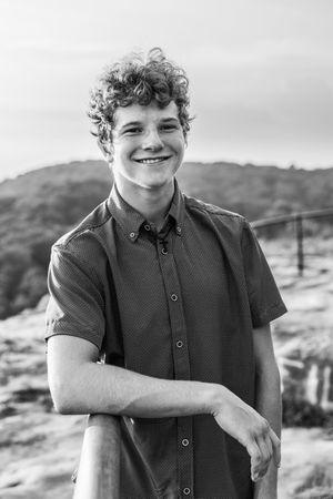Black and white portrait of a smiling person with curly hair wearing a button-down shirt outdoors in a natural setting.