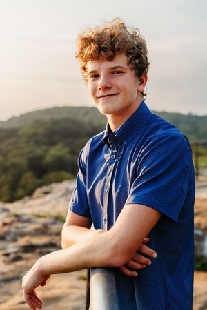 Young person in navy blue polo shirt smiling while leaning against railing with scenic mountain landscape at sunset.