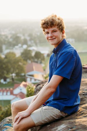 Someone in a blue polo shirt and khaki shorts sits on a rocky overlook with a blurred residential view in the background.