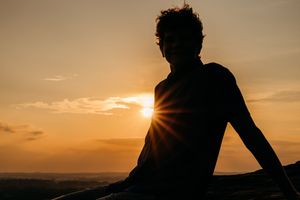 A silhouetted person sits on a hill against a dramatic orange sunset with sun rays peeking through clouds.