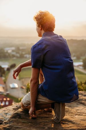Person in blue shirt sits on rocky overlook at sunset watching over valley vista and suburban landscape below.