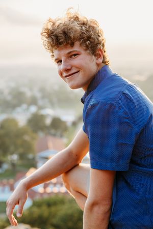 Someone with curly hair wearing a blue polo shirt smiles while posing outdoors during golden hour.