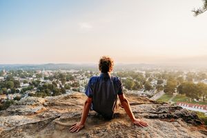 A person sits on a rocky cliff edge overlooking a scenic suburban town vista during golden hour.