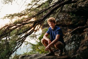 A hiker in a blue shirt crouches on a rocky outcrop at sunset with tree branches framing the scenic mountain view.
