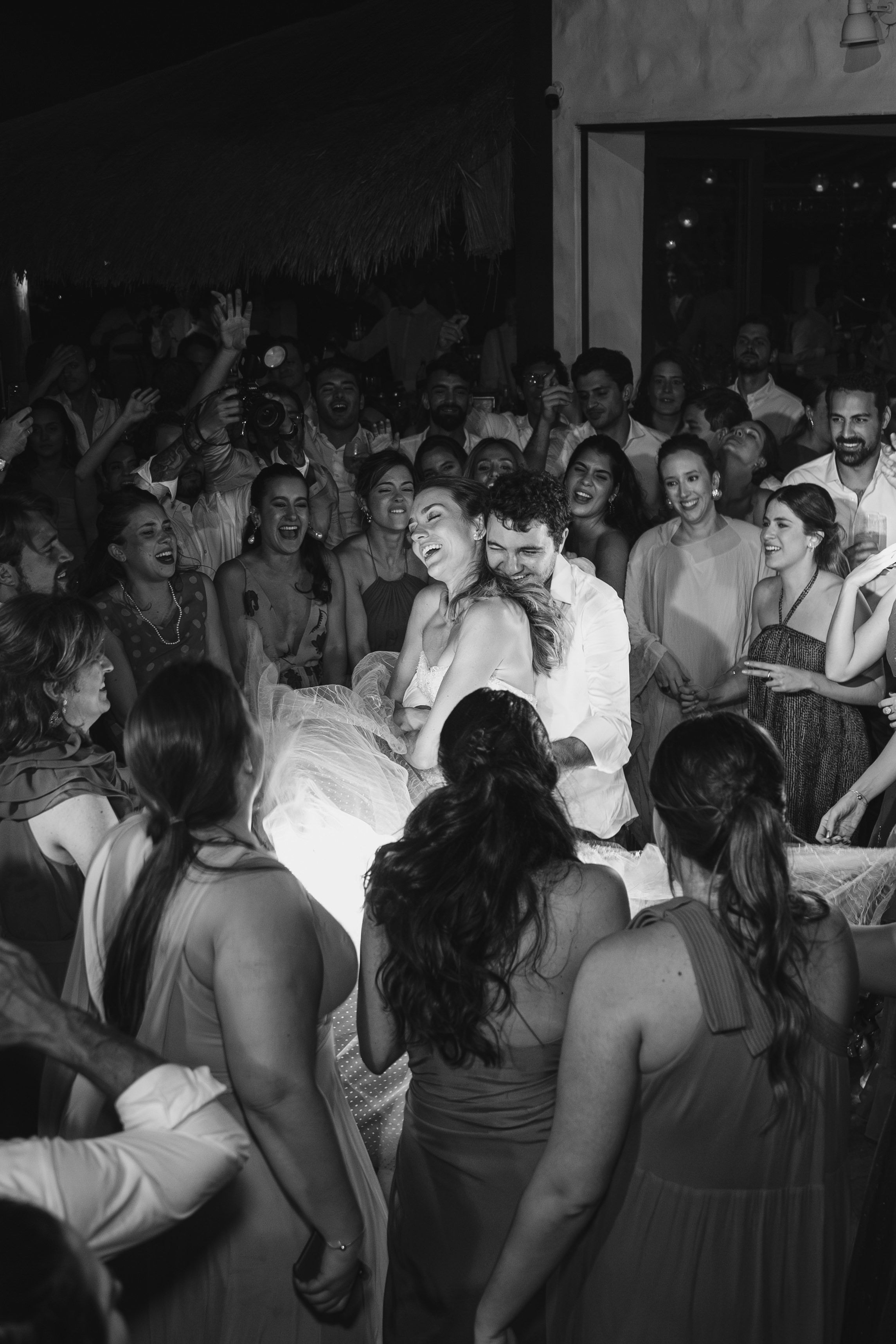 A crowd of people dancing and celebrating at what appears to be a wedding reception or party in a dimly lit venue.