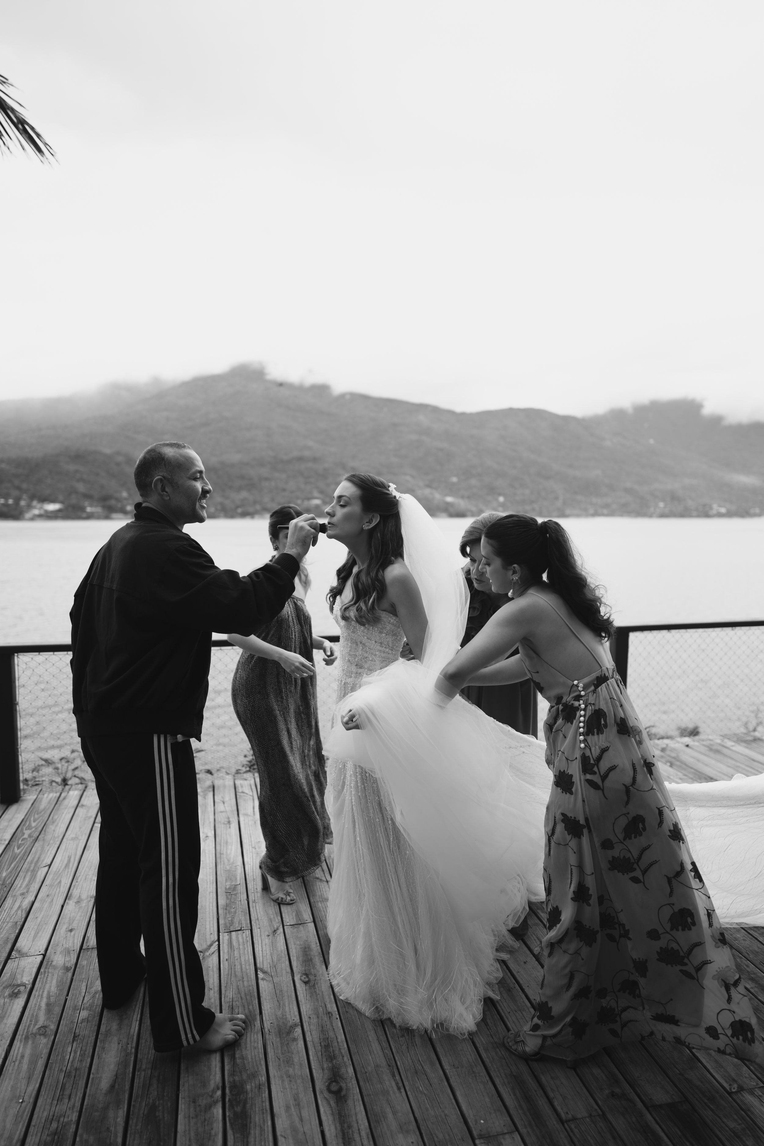 Wedding party enjoys a drink on a wooden deck overlooking a scenic mountain lake in black and white photo.