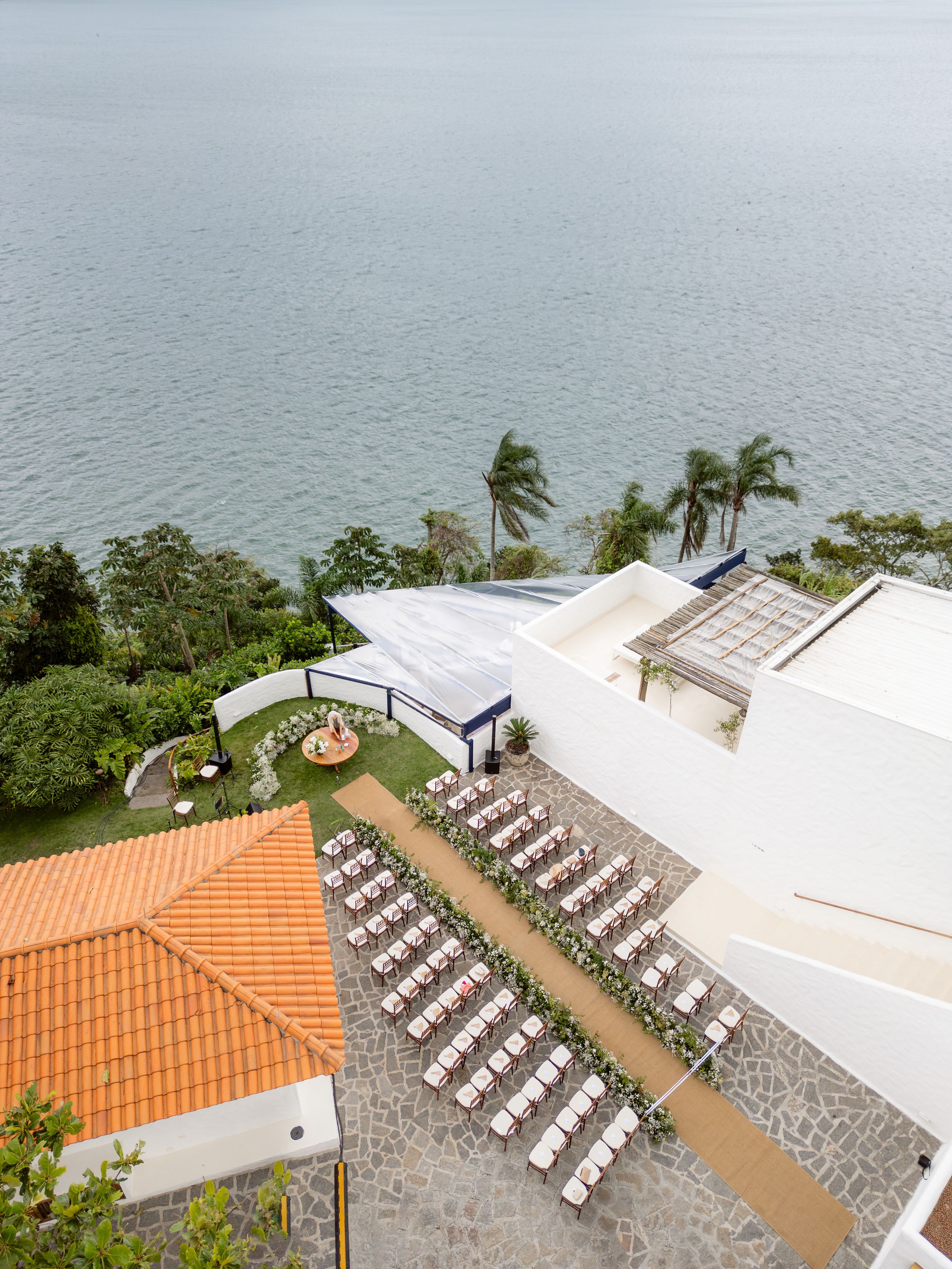 Aerial view of an outdoor wedding venue with white chairs arranged in rows overlooking the ocean and palm trees swaying in the wind.