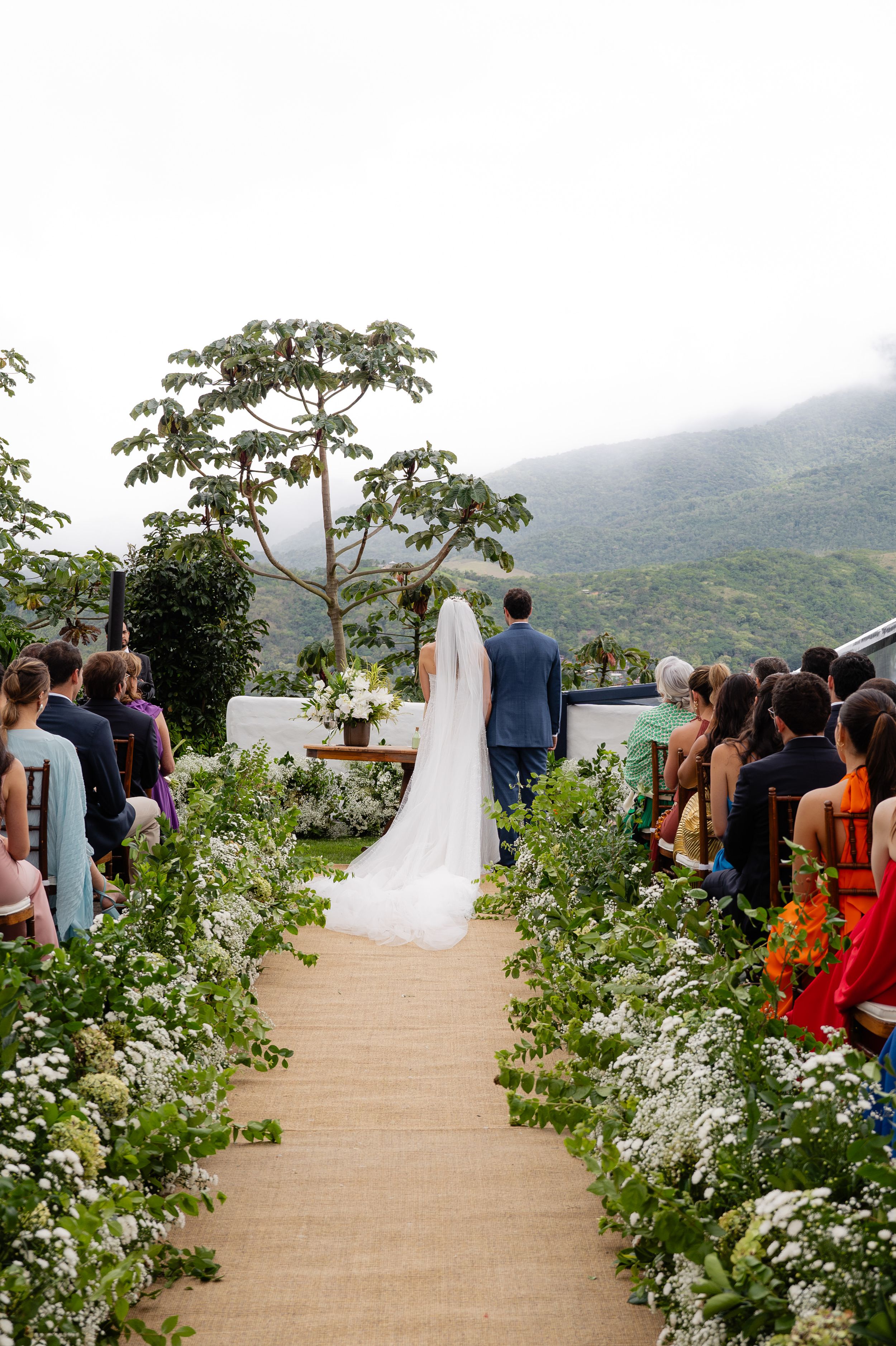 An outdoor mountain wedding ceremony set against misty hills with guests gathered on a decorated path.