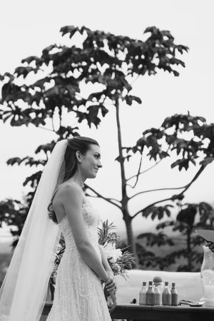 Black and white photo of a bride in lace dress standing beside a tree during an outdoor wedding celebration.