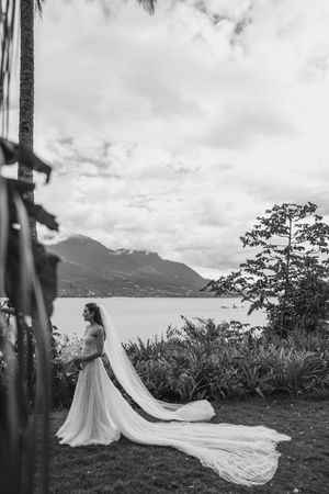 A bride in a flowing white gown with a long train stands at a scenic mountain lakeside overlook in black and white.