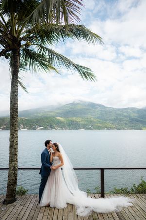 Couple in wedding attire embraces on a wooden deck overlooking a serene lake surrounded by mountains and palm trees.