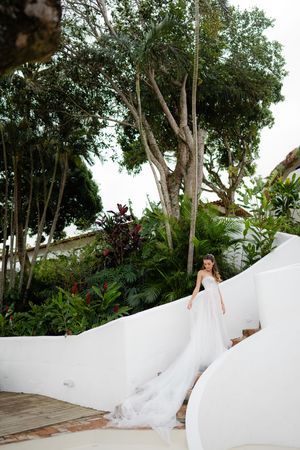 Elegant white wedding dress with long train flows down curved white architectural steps in a lush tropical garden setting.