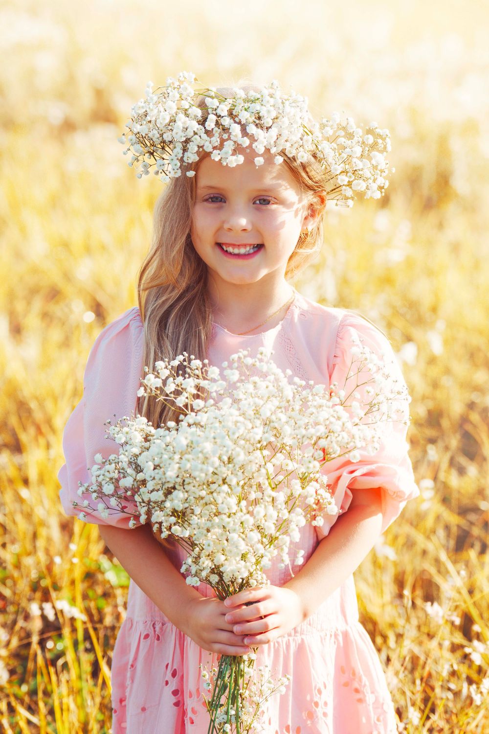 Young person in pink dress holding baby's breath flowers in sunny field wearing floral crown.